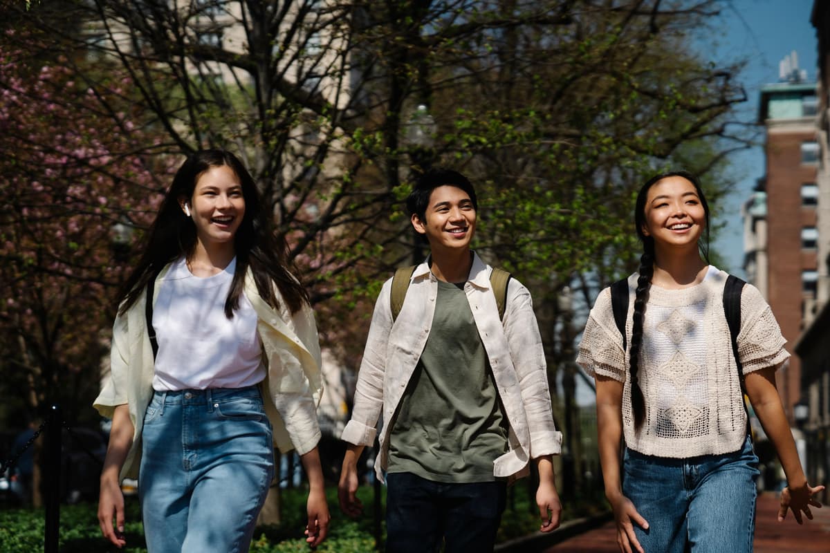 Three smiling students with backpacks walking together on a tree-lined university campus path.