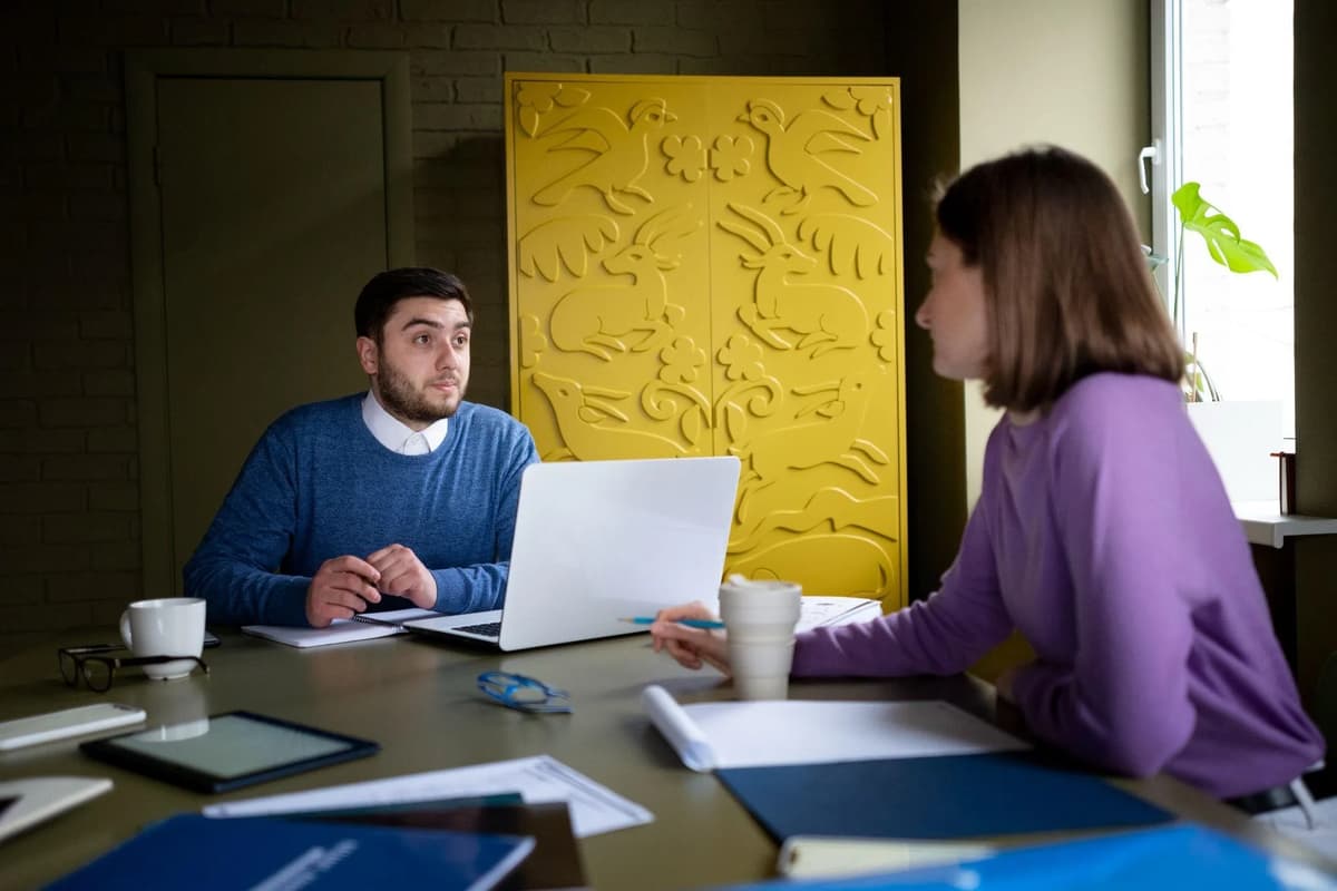Two professionals seated across a table with an open laptop, notebooks, and coffee cups.