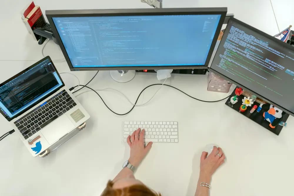 Software engineer working at a multi-monitor workstation, writing and debugging code on a laptop and external screens in a modern software development environment.