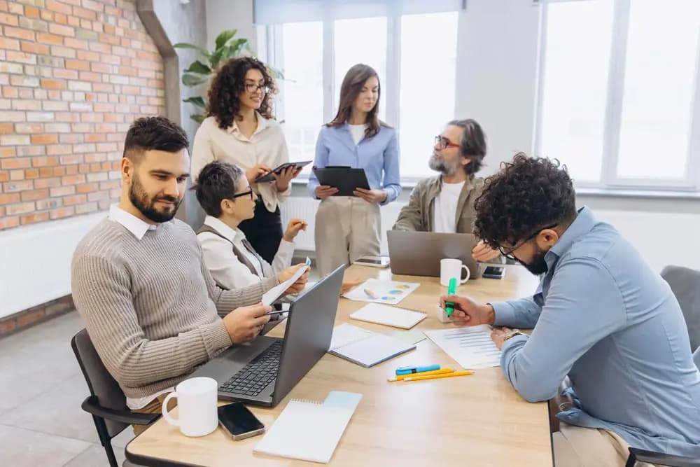 Business analytics team collaborating in modern office using laptops and AI-powered dashboards, demonstrating predictive data analysis and strategic decision intelligence in 2026