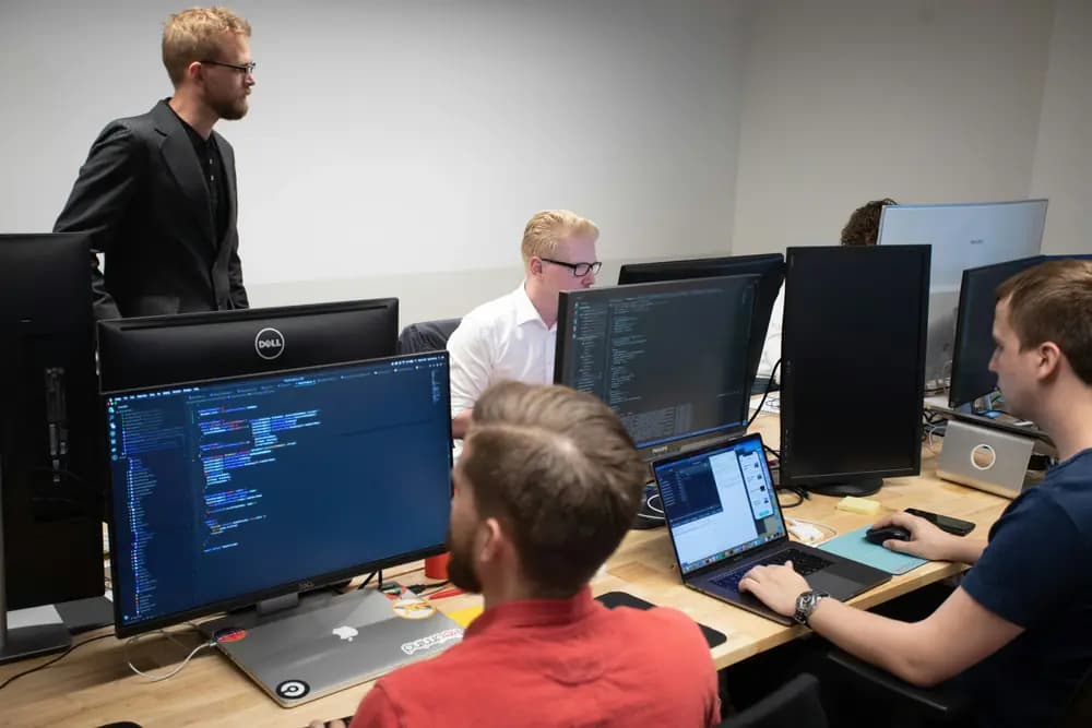 Backend engineers collaborating in an office environment, working on scalable software systems with multiple monitors displaying backend code, APIs, and system architecture.