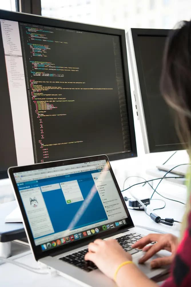 Software engineer working on a laptop with dual monitors displaying code in a modern development workspace