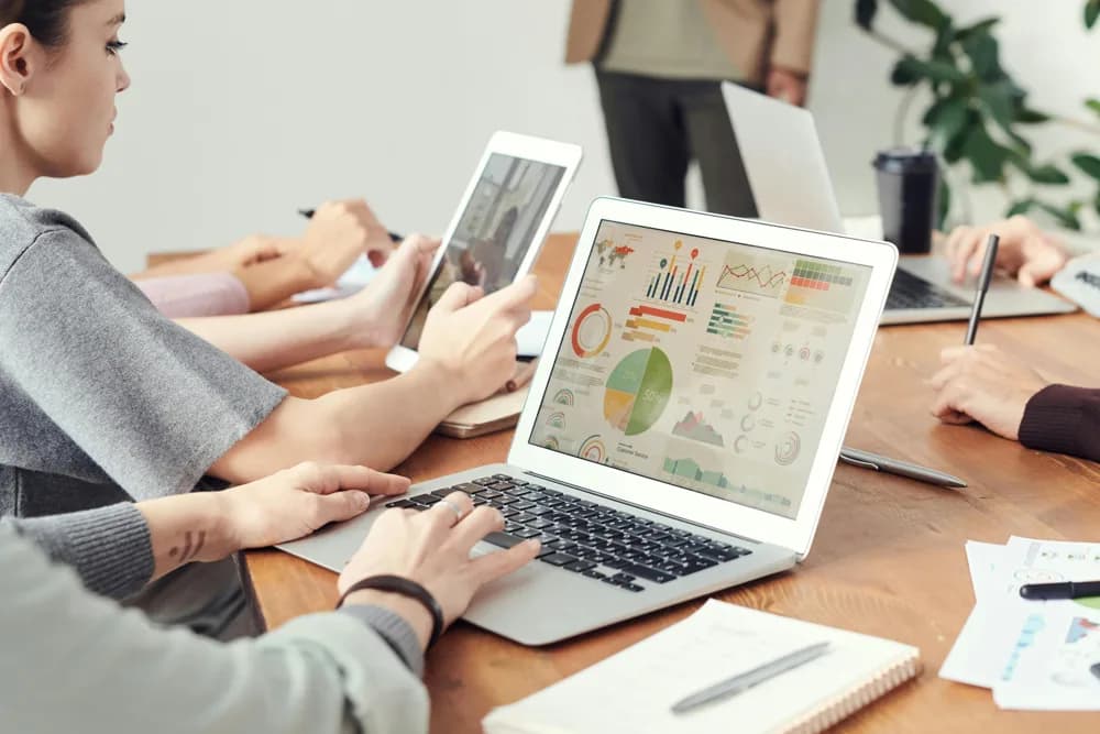 Team members working on laptops and tablets during a meeting, analyzing data dashboards, charts, and business analytics together.