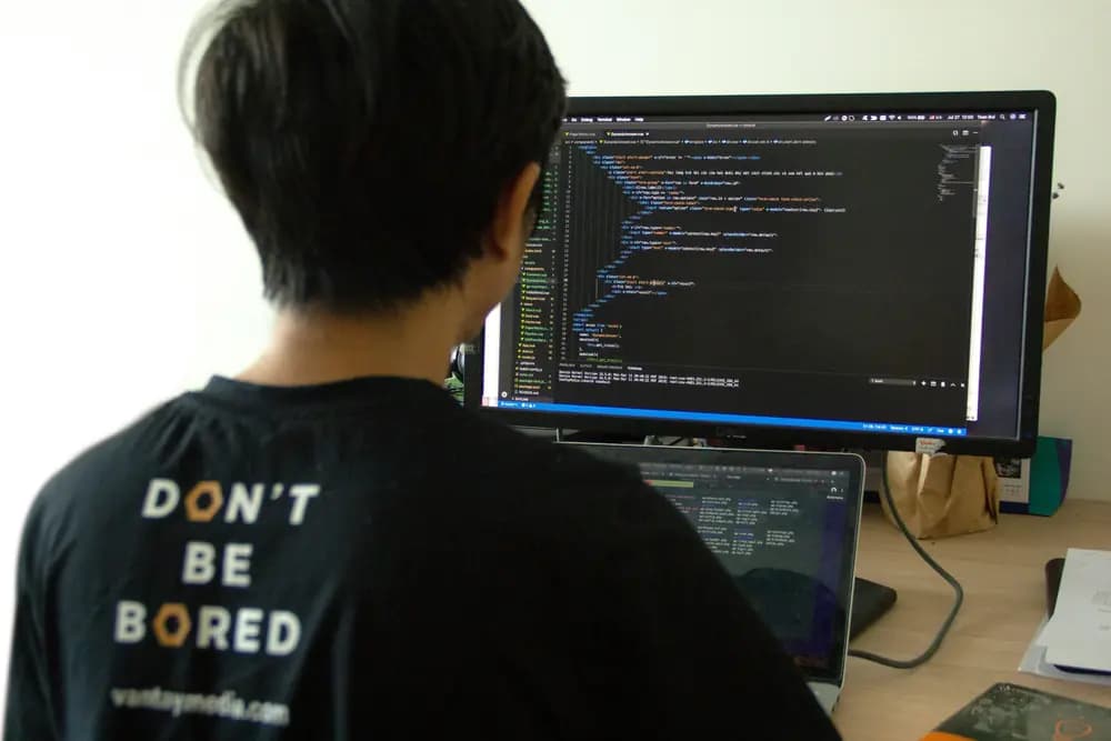 Software engineer writing and reviewing source code on a desktop monitor and laptop, working on web application development in a modern programming workspace.