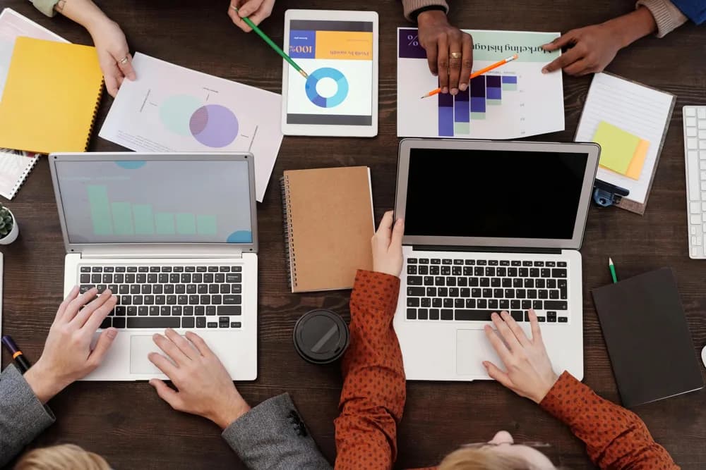 Top view of a team collaborating at a desk, working on laptops and tablets while reviewing charts, graphs, and data reports during a planning session.