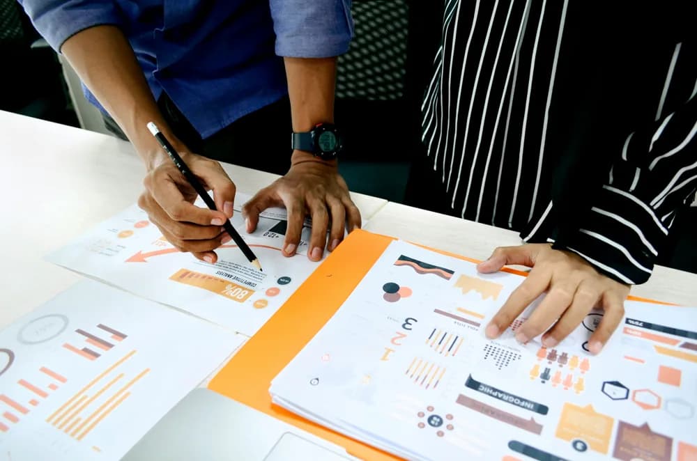Two colleagues collaborating at a desk, reviewing printed charts and data reports while one points to analytics with a pen.